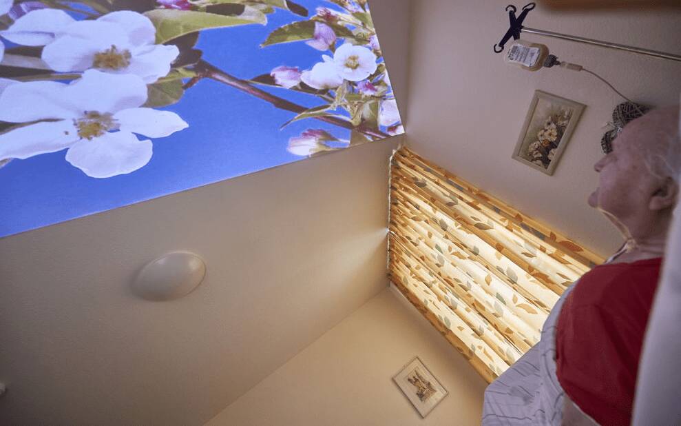 Eine bettlägerige Seniorin blickt auf ein an die Decke projiziertes Blumenbild.